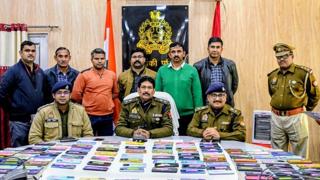 A group of police officers from Barabanki Police Department standing together in a formal event inside a conference room. They are holding smartphones that were recently recovered by the department's mobile recovery team. The background shows banners and insignias of the police force, with the Superintendent of Police presenting the phones to their respective owners. Smiles and gratitude are evident among the participants.